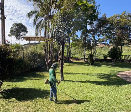 Roçada de gramado em área ampla, com árvores e céu aberto